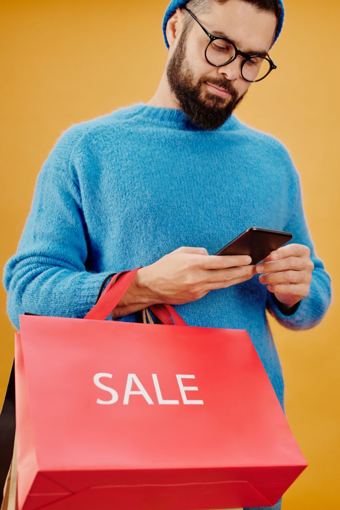 Bearded man holding a sale bag and smartphone in a studio setting.